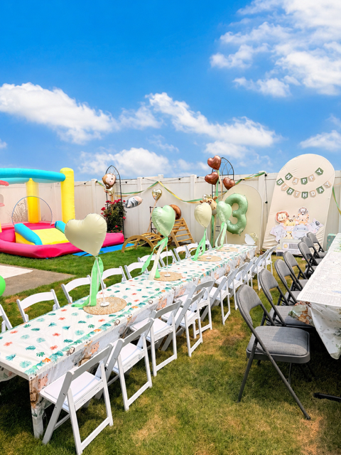Outdoor birthday party setup with decorated tables, balloons, and a bounce house, under a blue sky with some clouds.
