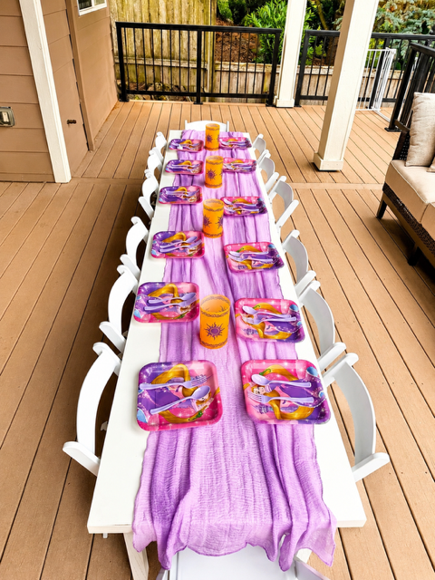 Outdoor dining table set for a children's party, with purple trays, orange cups, and yellow utensils, on a pink and purple table runner on a wooden deck.