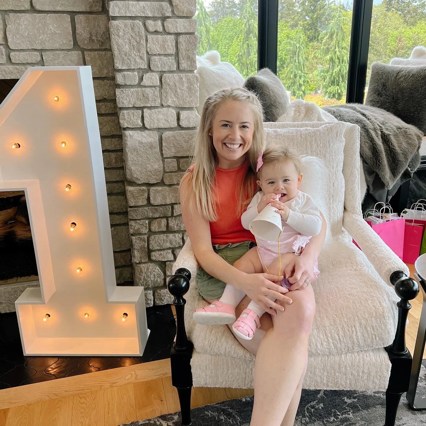 A woman and a young girl sitting on a beige armchair, smiling at the camera. The girl is holding a coffee cup and wearing pink shoes and a pink bow in her hair. There is a large illuminated number one letter and a stone fireplace in the background, along with a window showing green trees outside.