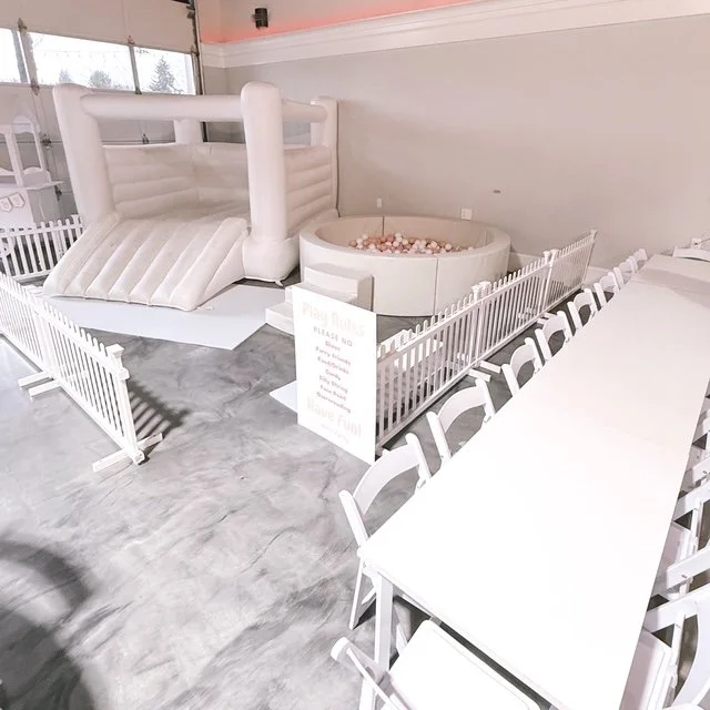 Indoor children's play area with an inflatable slide, small ball pit, and white fencing, with white tables and chairs nearby.