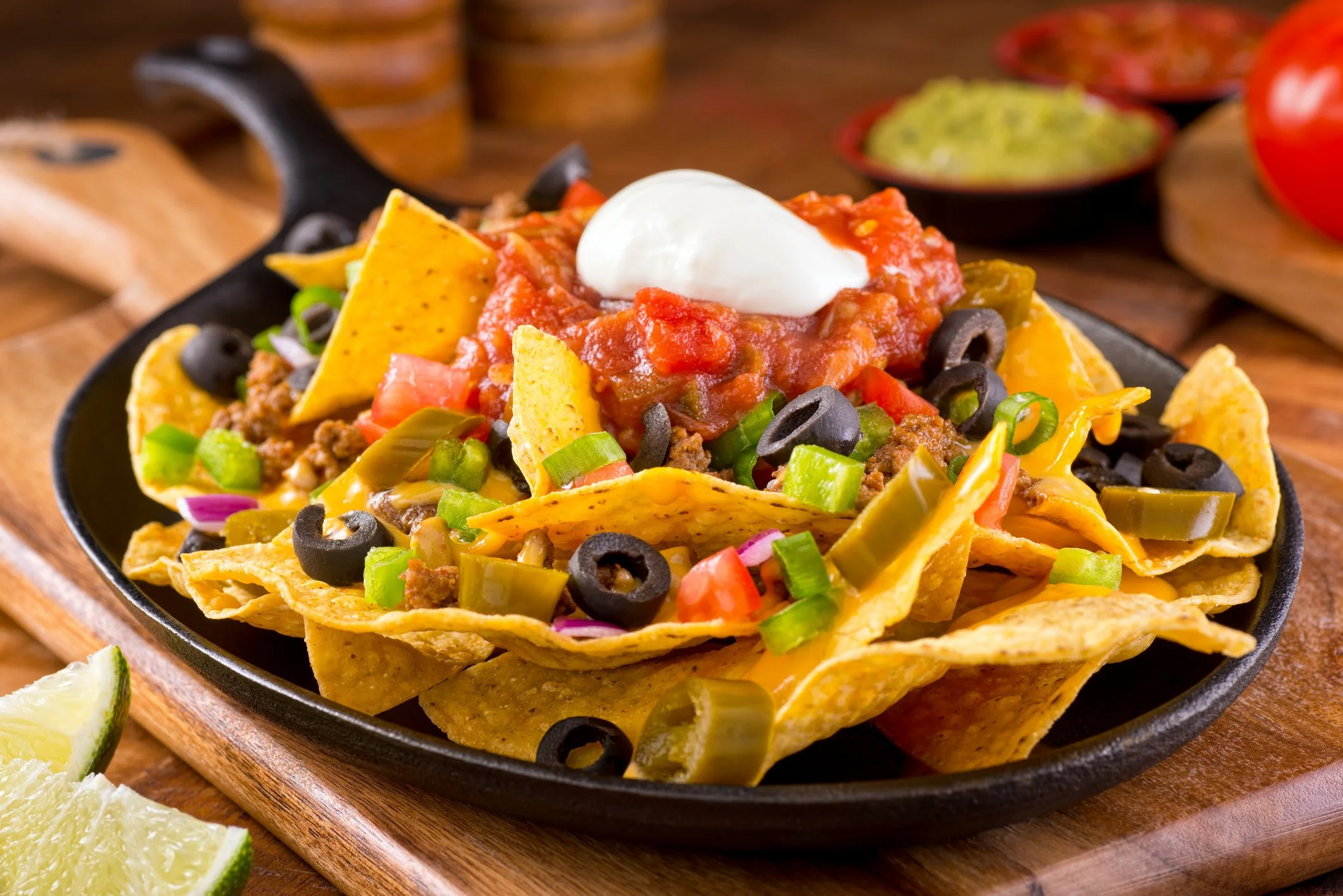 Plate of nachos with cheddar cheese, ground beef, black olives, jalapeños, diced tomatoes, green onions, salsa, and a dollop of sour cream.