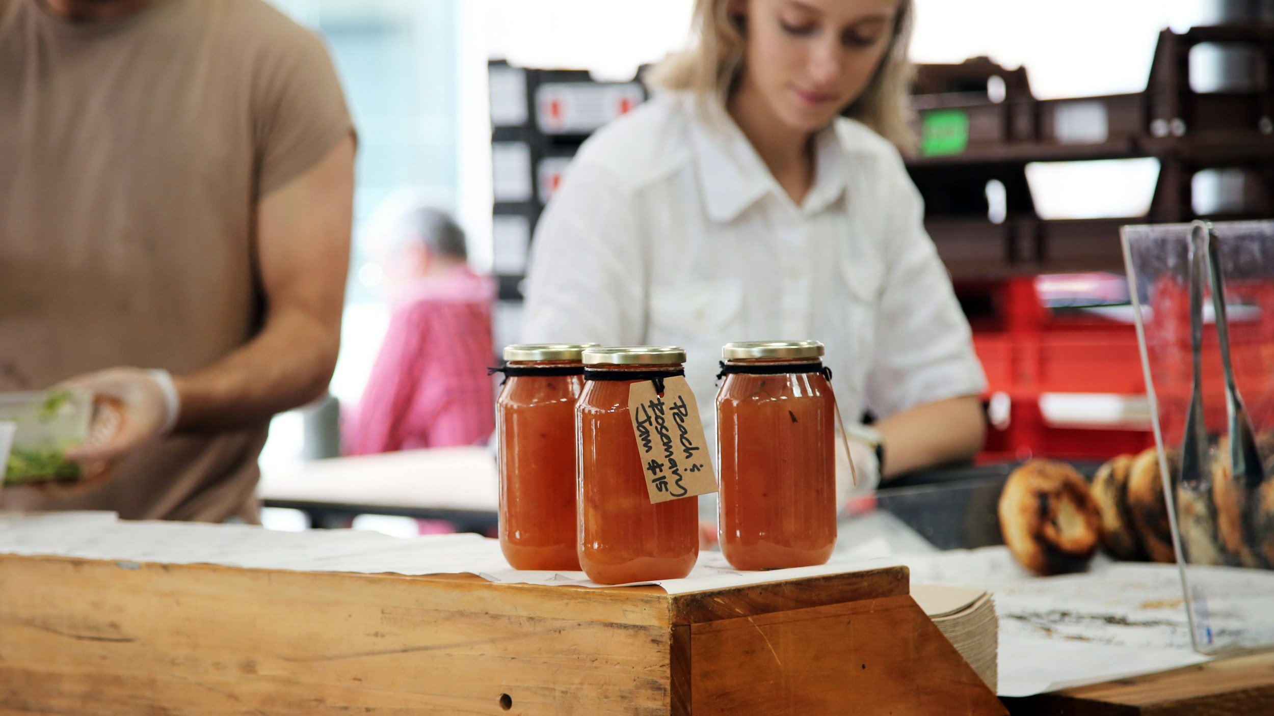 Three jars of peach and rosemary jam displayed on a wooden counter at a market, with a person in the background preparing food.