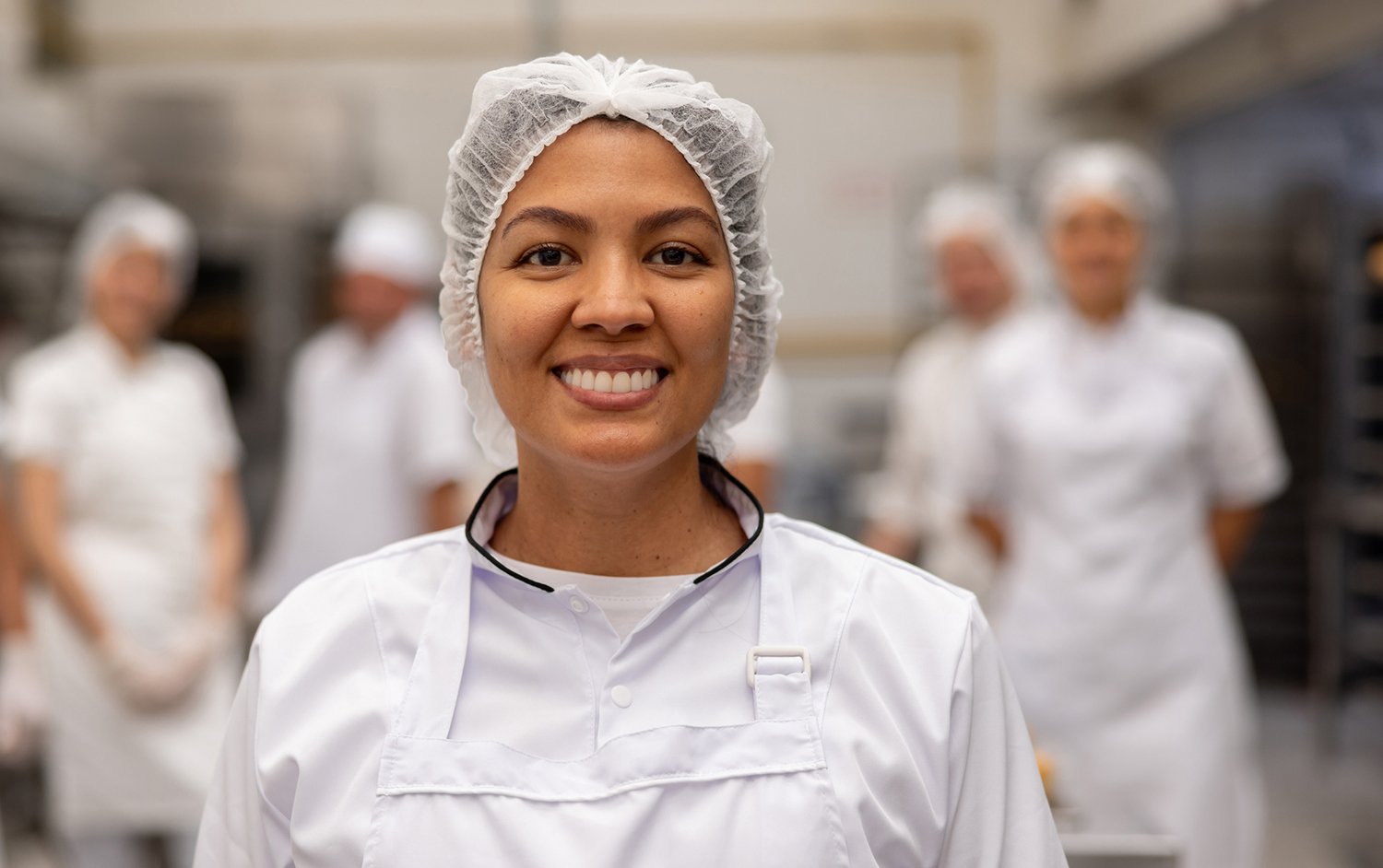 Smiling chef wearing a hairnet and white uniform in a commercial kitchen, with blurred colleagues in the background.