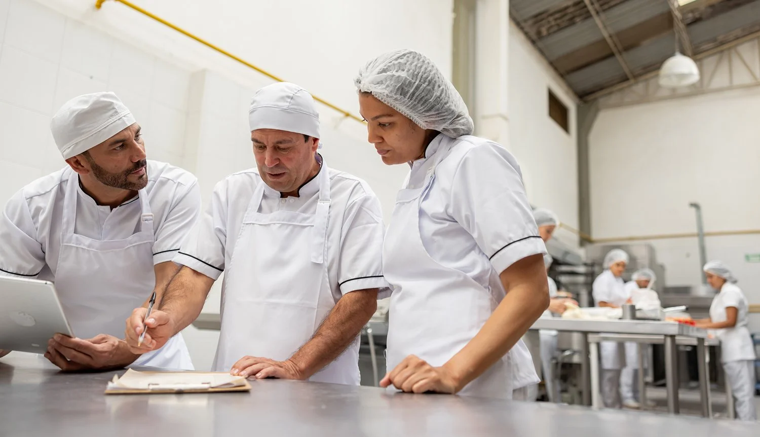 Group of bakers wearing white uniforms and hairnets discussing in a commercial kitchen.