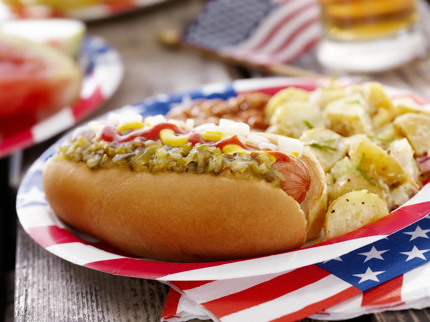 Close-up of a hot dog with condiments and potato salad on a patriotic paper plate with an American flag design.