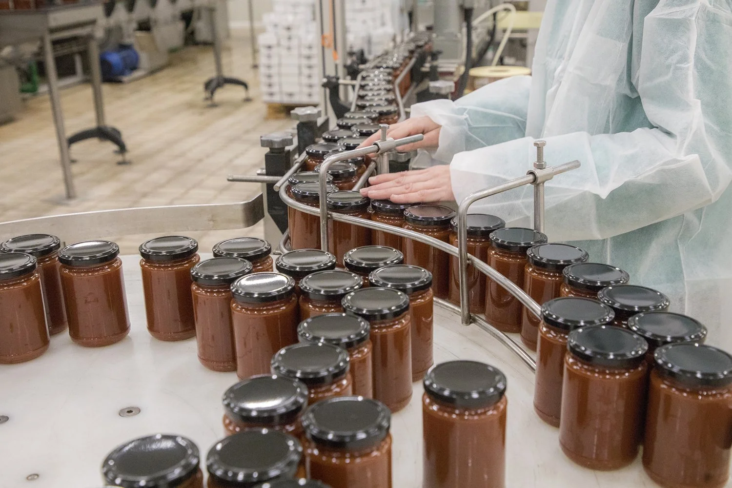 Person inspecting jars of sauce on a conveyor belt in a food factory.