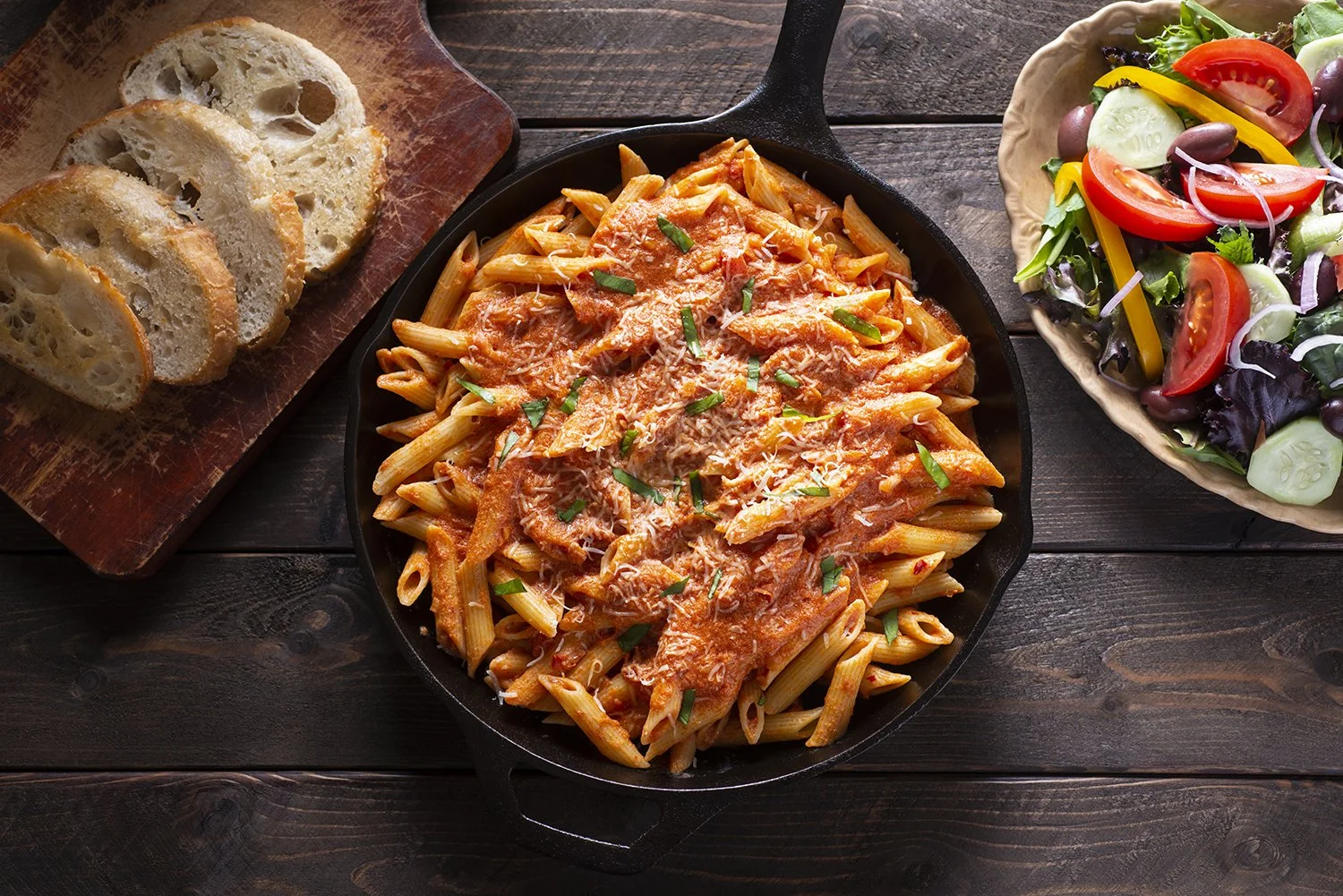 Penne pasta with tomato sauce and cheese in a skillet, sliced bread on a cutting board, and a mixed salad with tomatoes, cucumbers, olives, and peppers in a bowl.