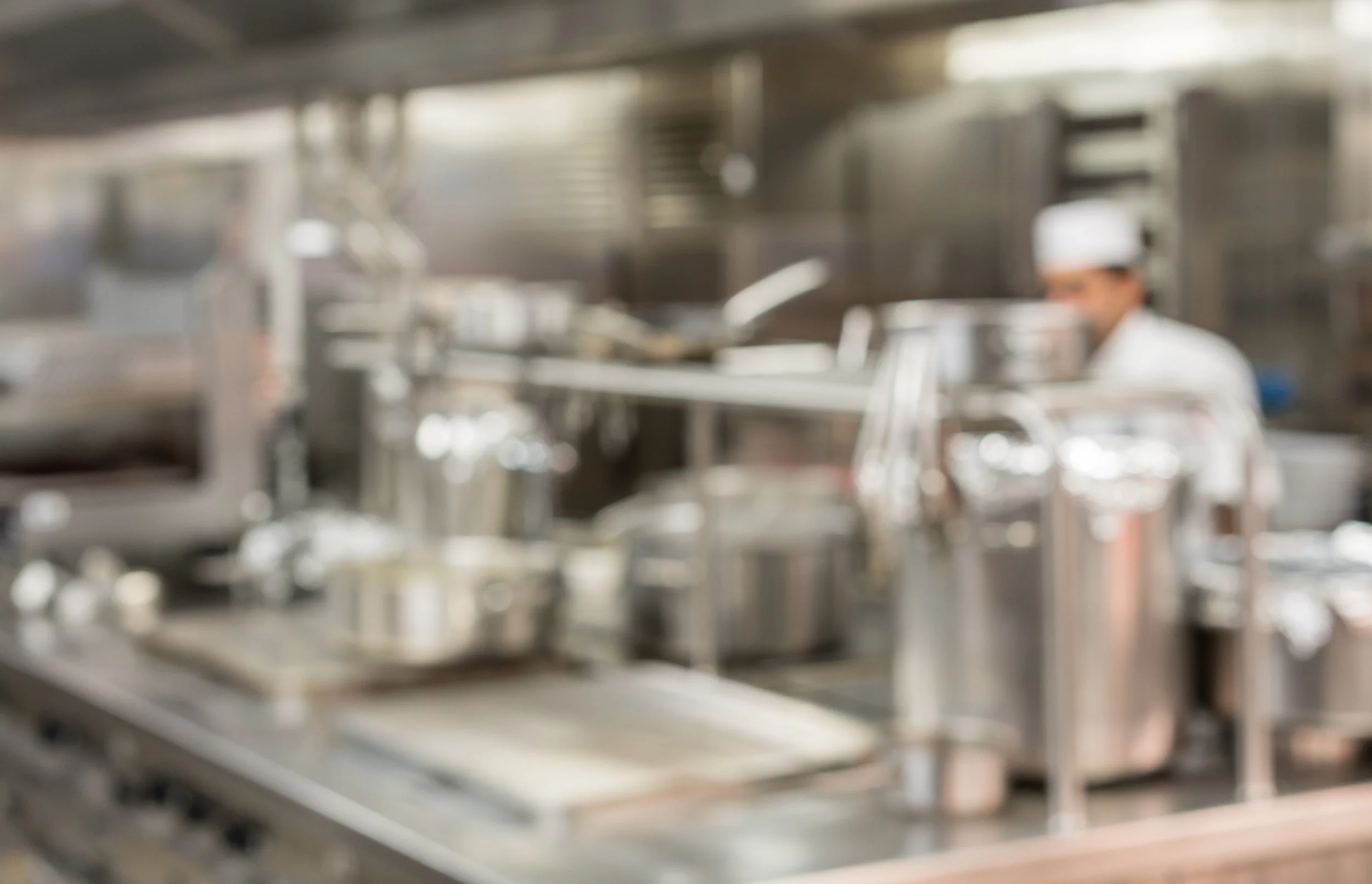 Blurred image of a commercial kitchen with stainless steel equipment and a chef in the background.