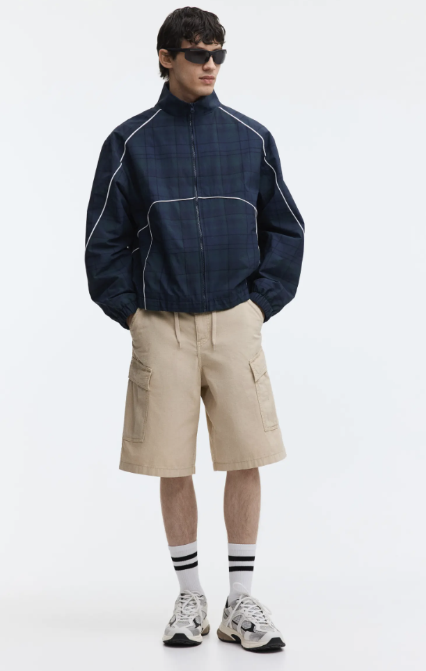 A man wearing tan-colored elevated cargo shorts with flat pockets, paired with a navy blue long-sleeve top and sneakers, showcasing a tailored summer style for 2026.