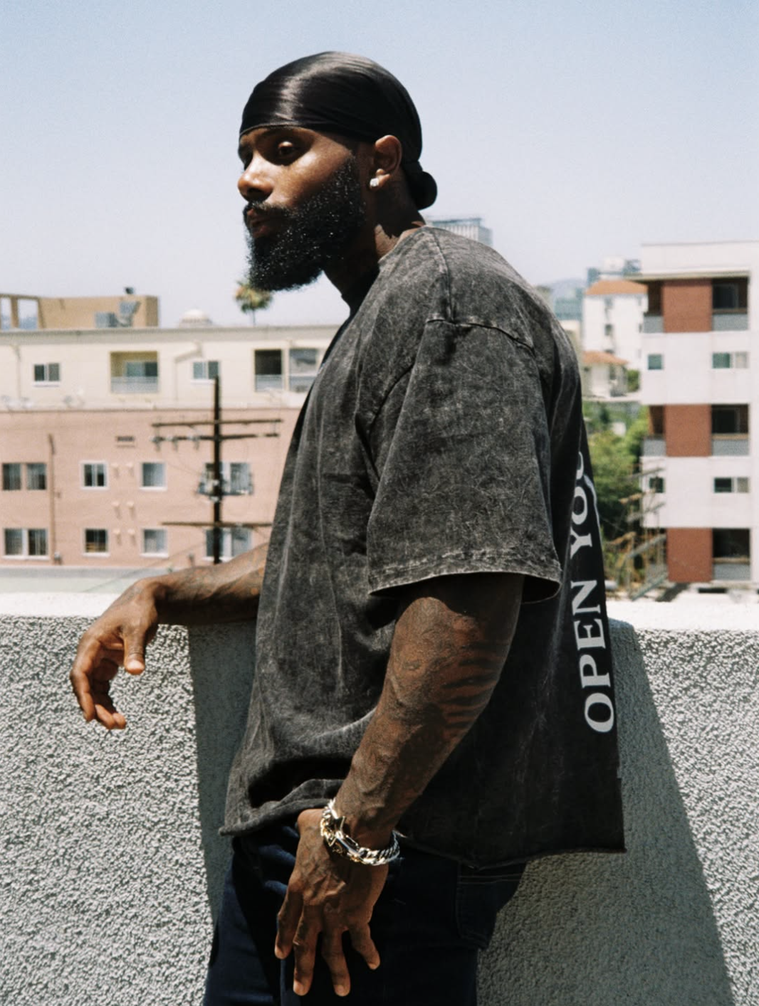 A man from the side wearing a black acid-wash graphic t-shirt with text on the back, silver chain bracelets, and tattoos on his arm, posing outdoors against a concrete wall.