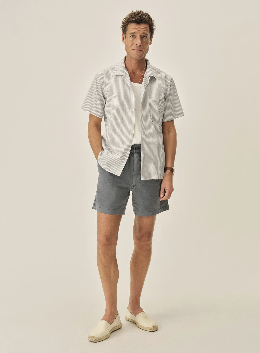 A man wearing khaki Buck Mason Deck Shorts paired with white canvas espadrilles, showing a rugged yet polished outdoor summer style.