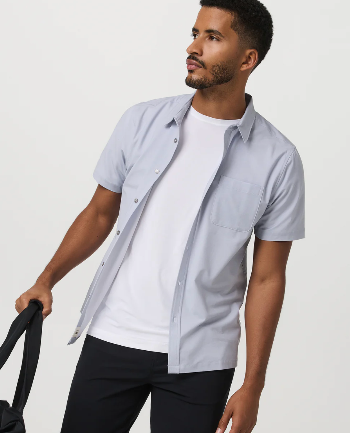 A side-angle shot of a man wearing a light blue short-sleeve Vuori Atmosphere linen shirt open over a classic white t-shirt, paired with black trousers.