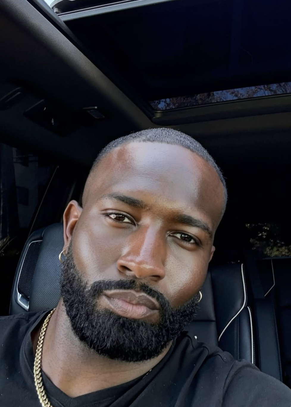 Close-up selfie of a man with a perfectly groomed short boxed beard, a clean buzz cut, and a gold chain, sitting inside a car.