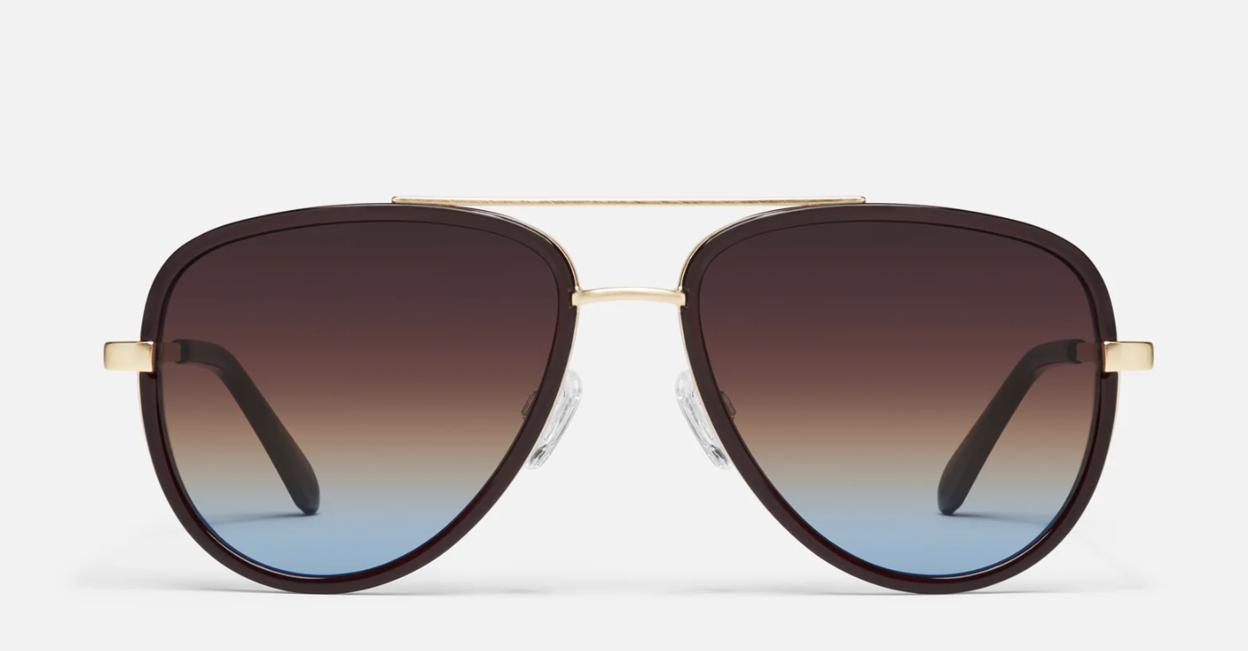 A close-up studio shot of Quay Australia All In aviator-style sunglasses, featuring a bold brown frame with a sleek gold double brow bar and gradient lenses fading from brown to a cool blue tint.