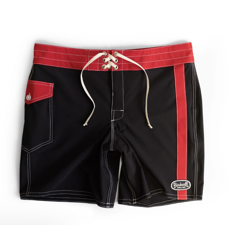 A close-up view of black Birdwell board shorts featuring a classic lace-up contrast cream drawstring waistband, a red side stripe, a buttoned red hip pocket, and the iconic Birdwell patch on the lower hem.
