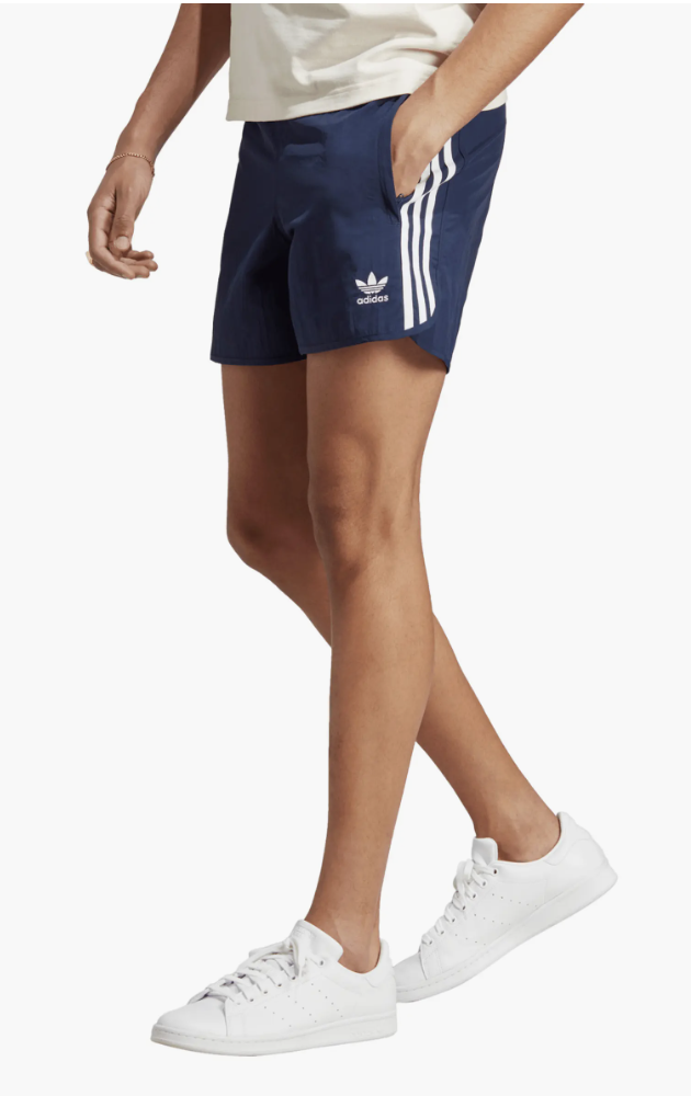 A man wearing navy blue adidas nylon sprinter shorts with white stripes, paired with classic white adidas Stan Smith sneakers.