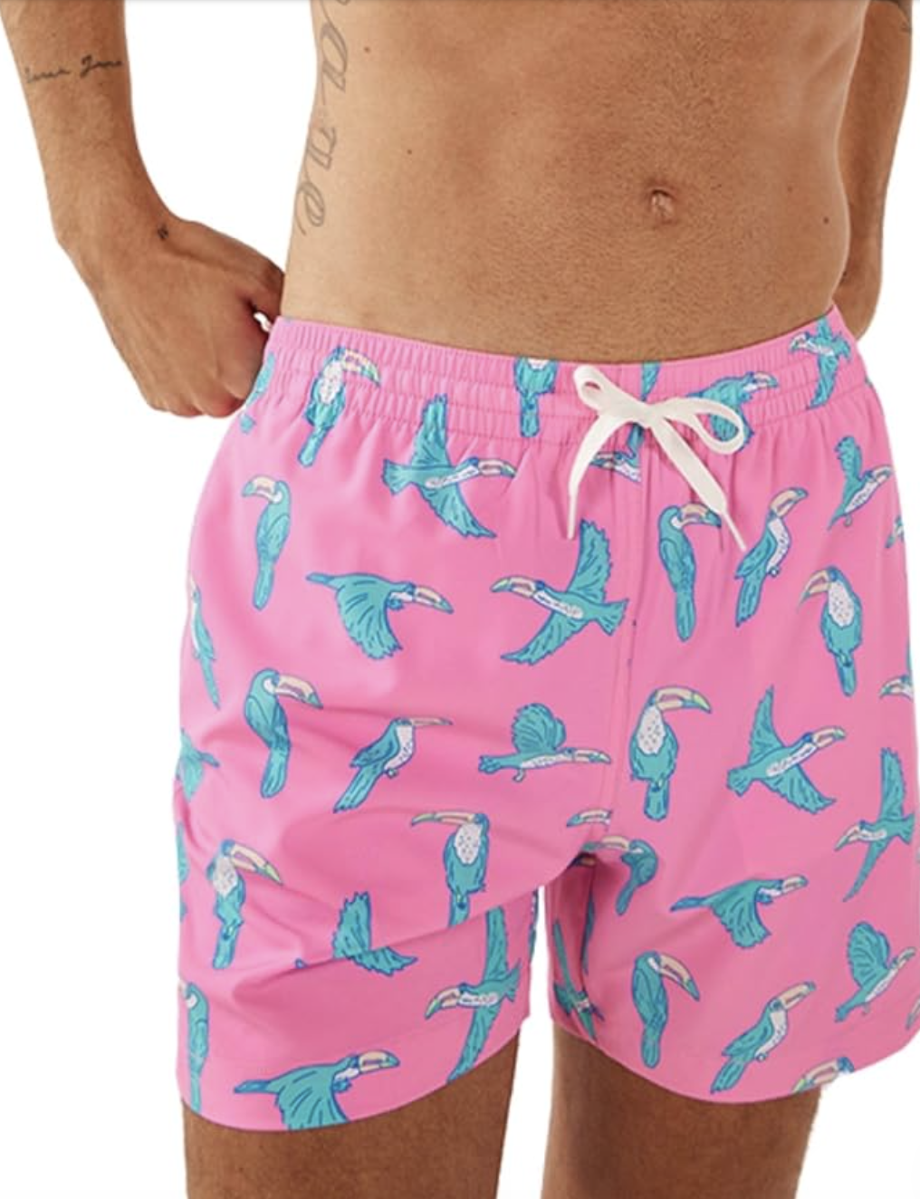 A close-up of a person wearing Chubbies The Everetts 5.5-inch inseam swim trunks featuring a vibrant pink background with a bright blue toucan pattern.