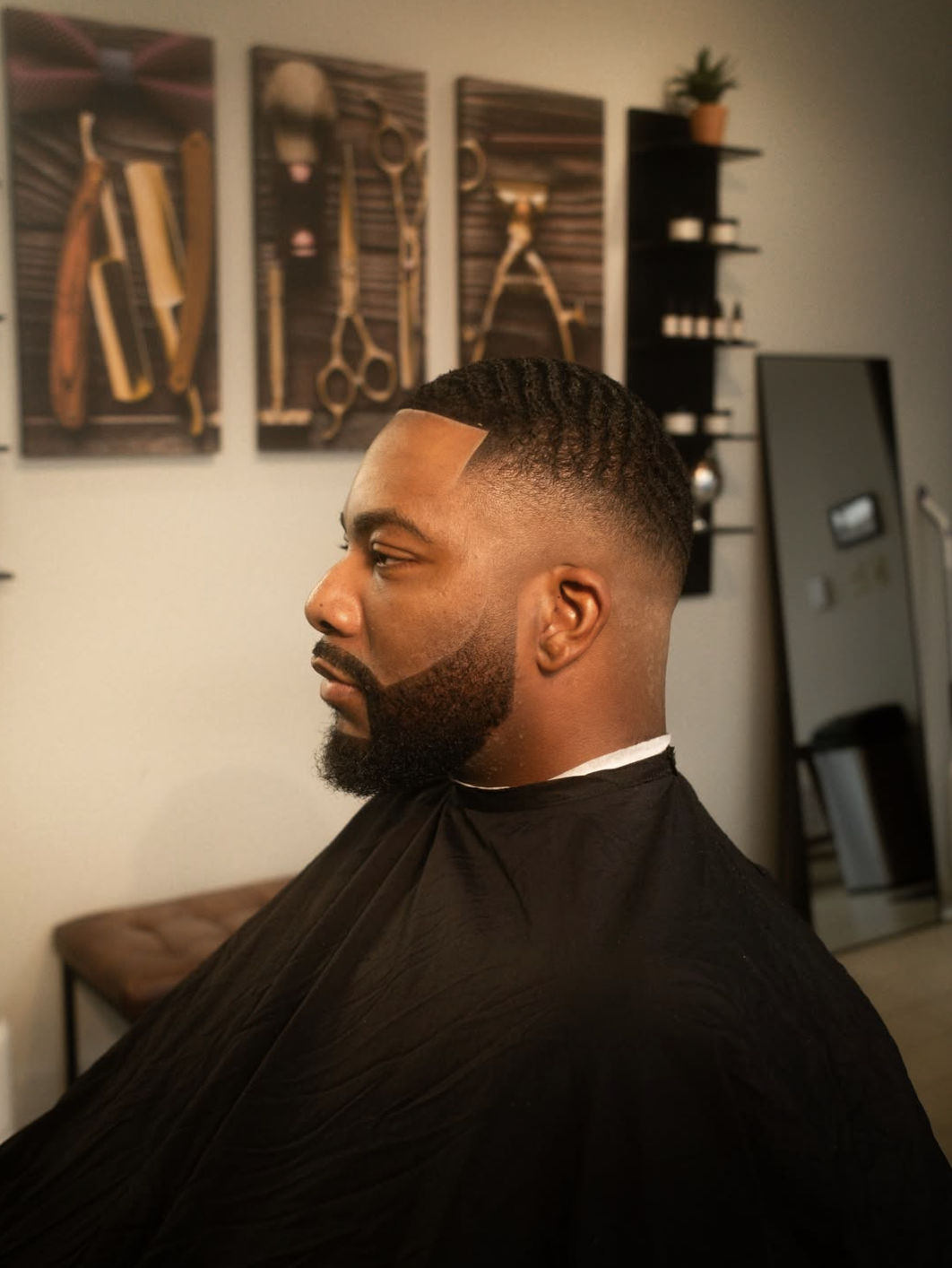 Side profile of a man with a perfectly lined short boxed beard, a clean mid-skin fade haircut, and crisp hair design lines, posing inside a barber shop with vintage grooming tools on the wall.