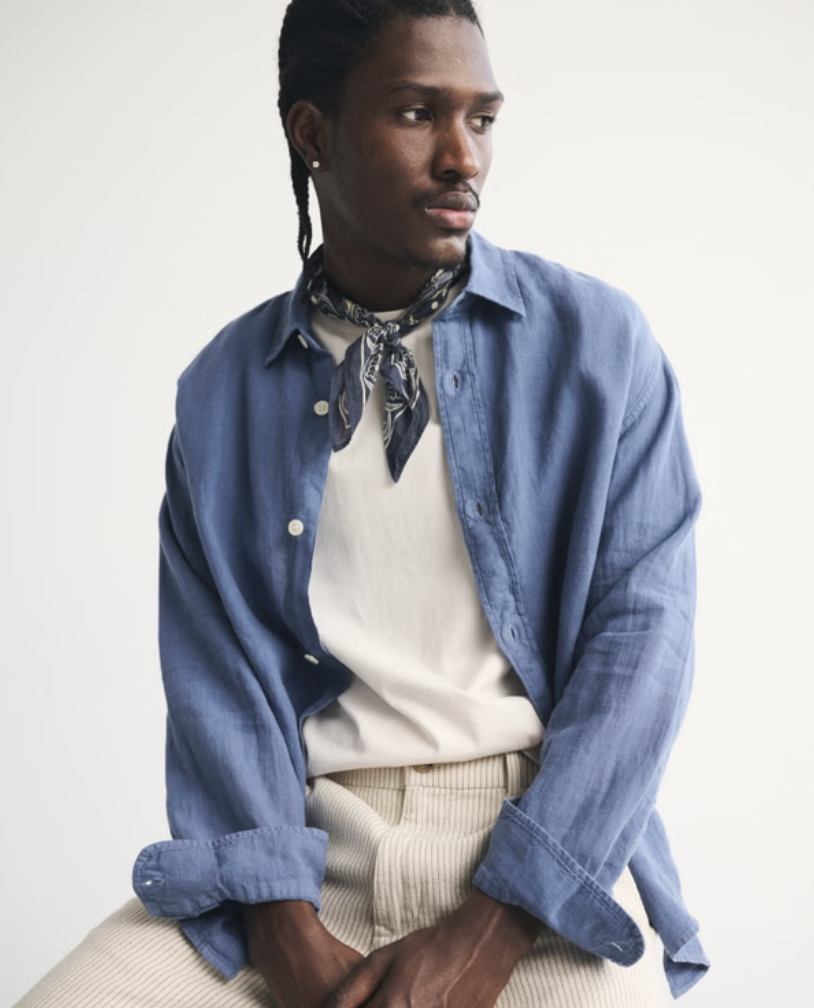 A man wearing an oversized light blue linen button-down shirt layered over a white t-shirt and cream trousers, styled for a relaxed 2026 summer look.