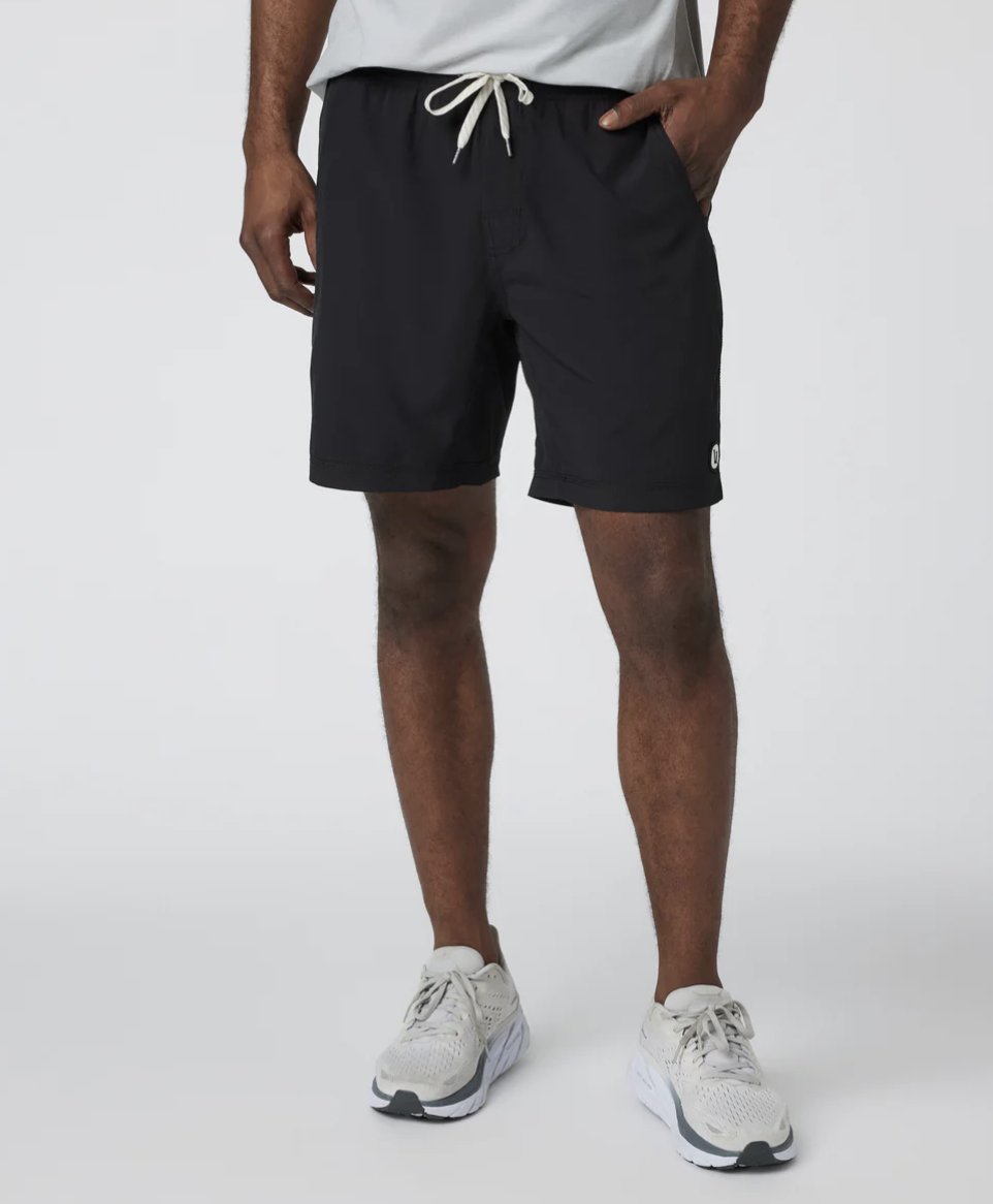 A man wearing black Vuori Kore 7-inch unlined shorts paired with white athletic sneakers, showing a versatile gym-to-street fit.