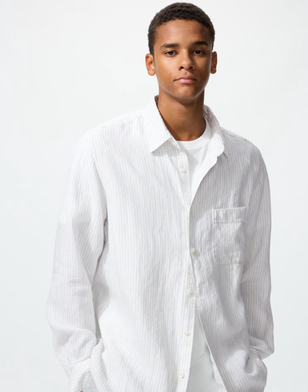 A close-up shot of a man wearing a Uniqlo Premium Linen long-sleeve shirt in a light white and blue vertical stripe pattern over a white t-shirt.