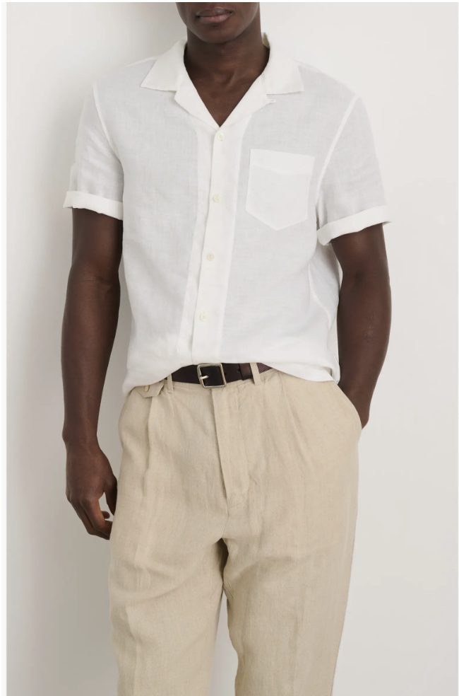 A close-up shot of a man wearing a classic white Alex Mill camp-collar linen shirt, styled tucked into pleated beige trousers with a dark brown leather belt.