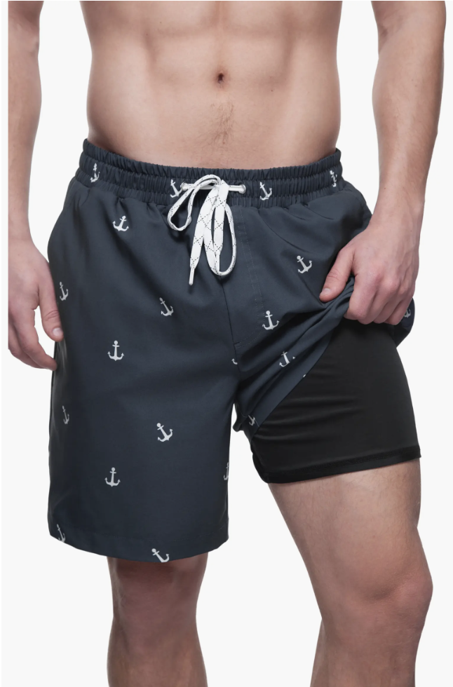 A close-up of a person wearing dark navy blue men's swim trunks featuring a white anchor print pattern and a supportive, black built-in compression liner.