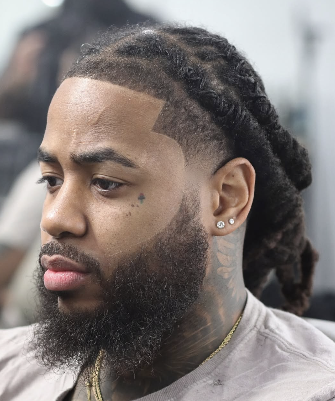 Side profile of a man with a sharp ducktail beard, a precise mid-skin fade haircut, and neat braided hair with crisp lineup edges, showcasing a tiny cross tattoo under his eye.