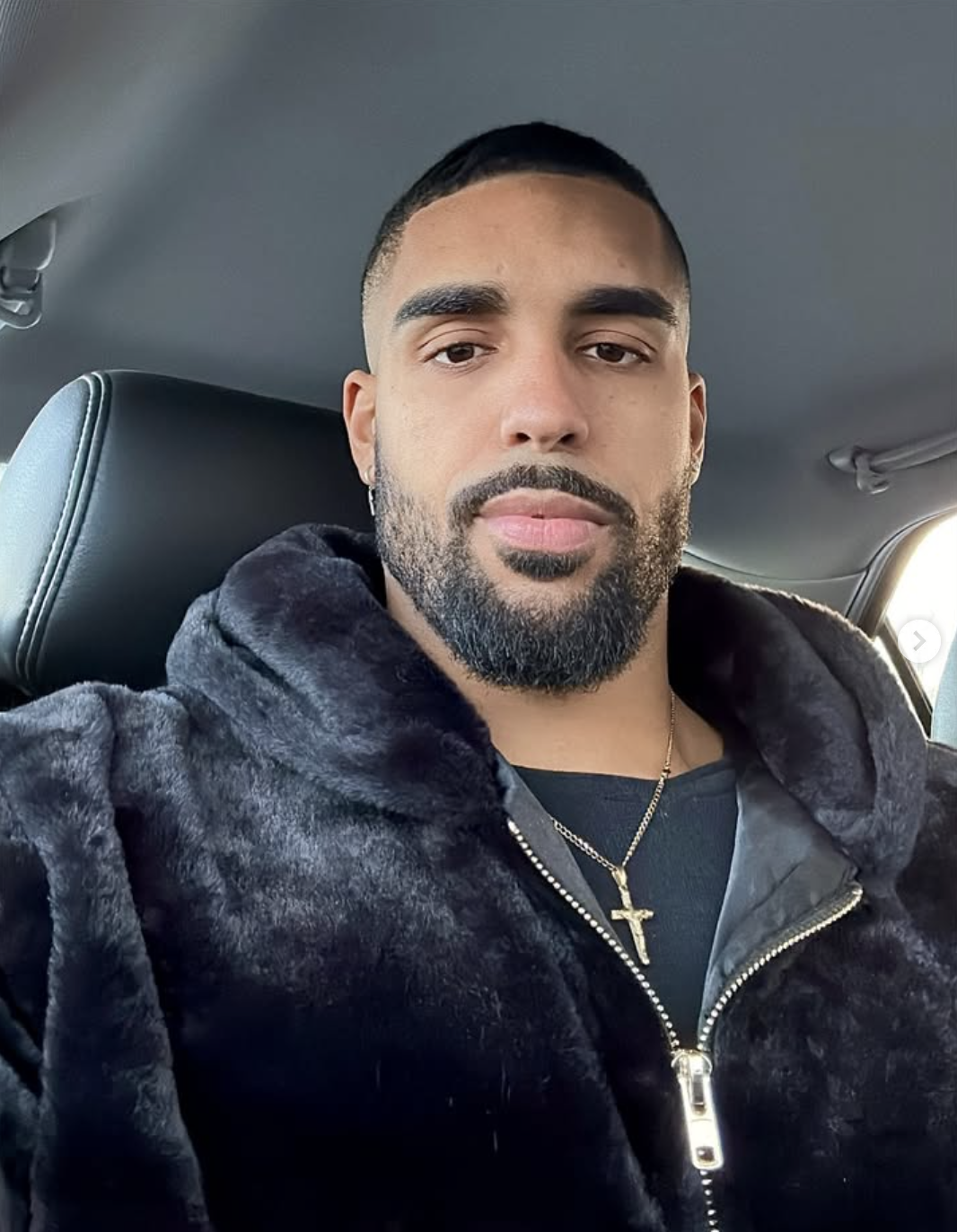 Close-up selfie of a man showcasing a clean 5 o'clock shadow light stubble beard and a fresh buzz cut lineup, wearing a black plush jacket and a gold cross necklace while sitting in a car.