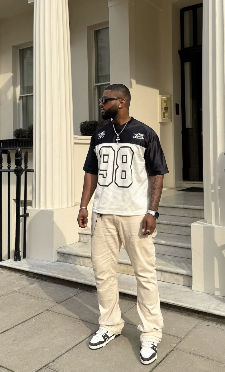A close-up view of a man standing on outdoor steps wearing a black and white short-sleeve sports jersey with the number 98, styled with light beige relaxed-fit cargo pants, sunglasses, and a silver watch.