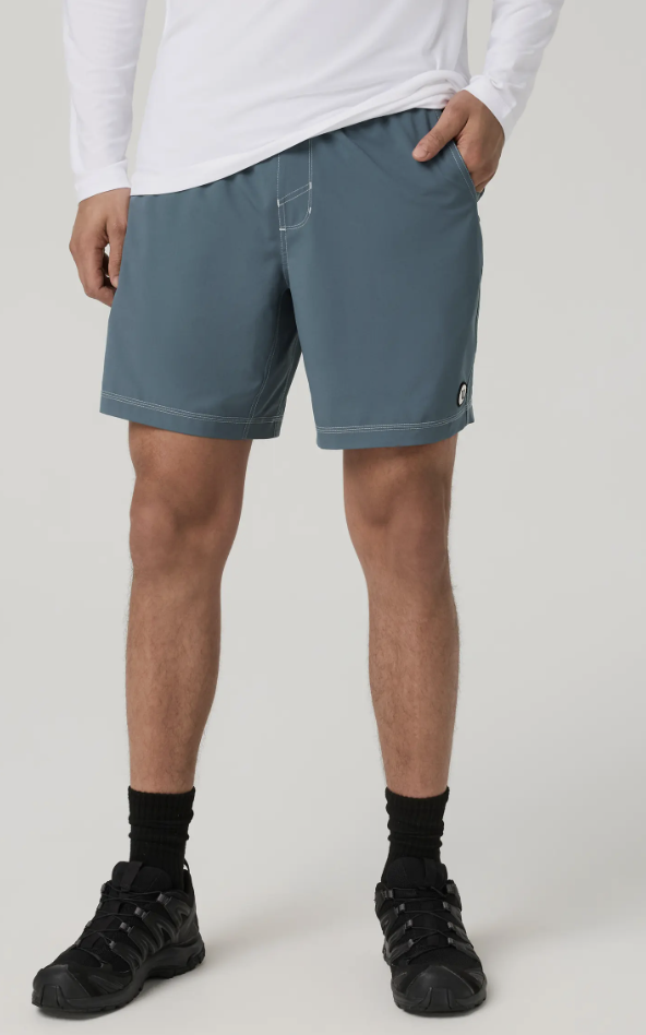 A close-up of a person wearing the steel blue Vuori Kore Volley Short paired with a white long-sleeve shirt, highlighting the clean fit, drawstring waist, and minimal brand logo on the hem.