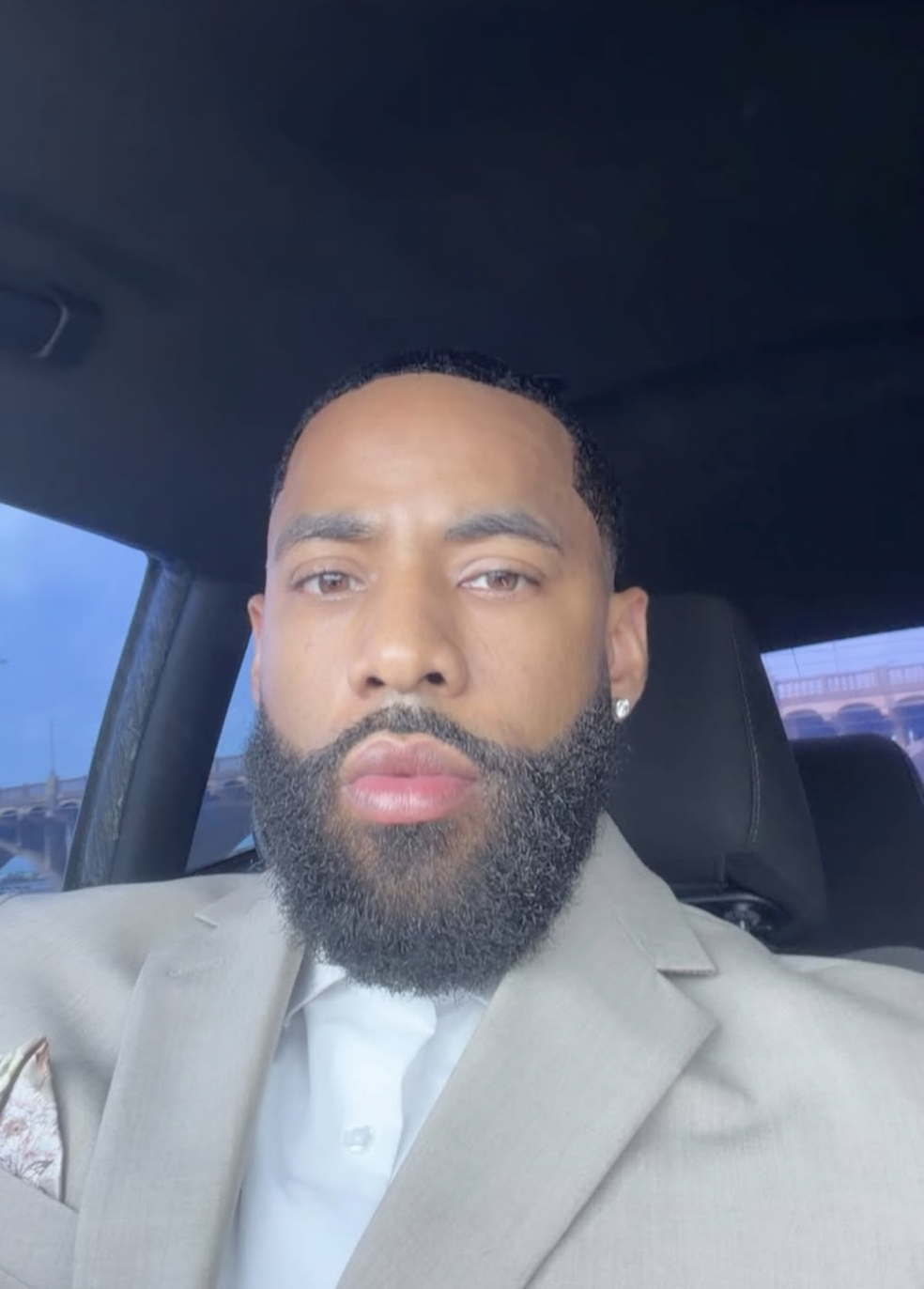 Front view portrait of a man with a perfectly lined short boxed beard and a fresh buzz cut, wearing a light beige suit jacket and a white dress shirt while sitting inside a car.