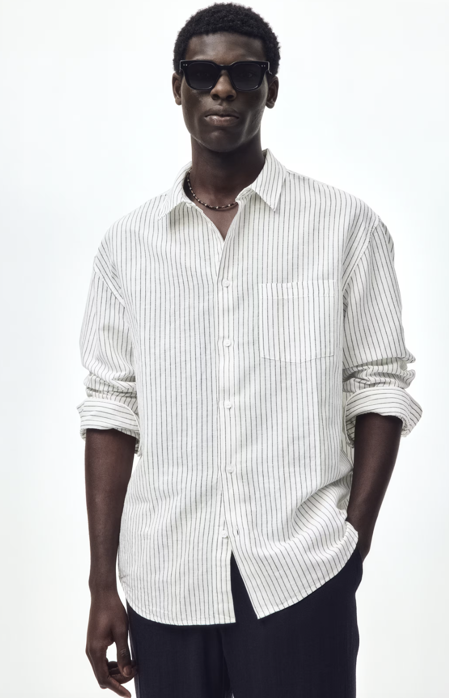 A close-up shot of a man wearing an H&M Regular Fit linen shirt with fine vertical grey stripes, accessorized with a dark beaded necklace and black sunglasses.