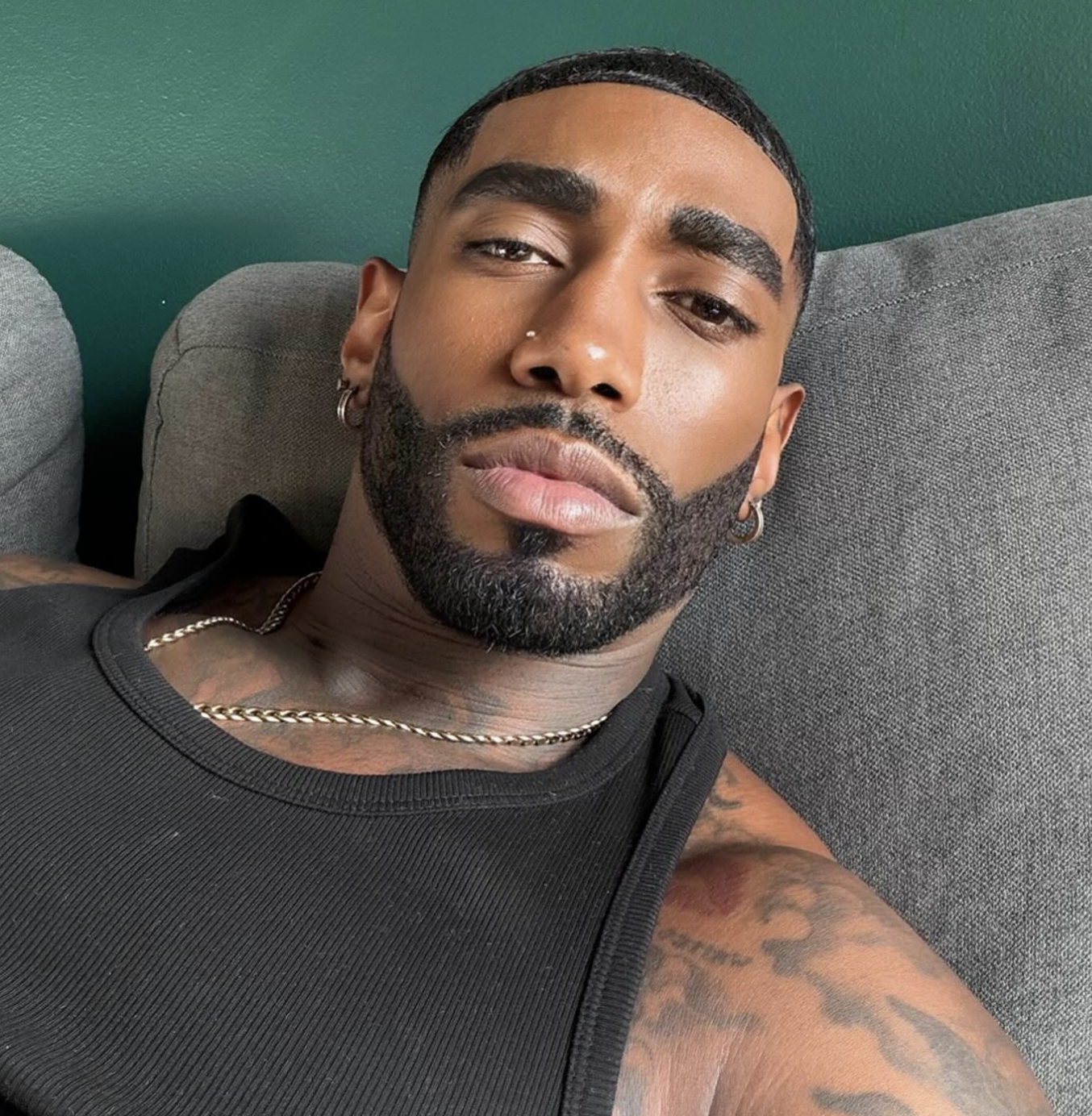 Close-up portrait of a man with a completely clean-shaven face and a sharp mid-skin fade haircut with textured waves, wearing a black tank top and a gold chain necklace.