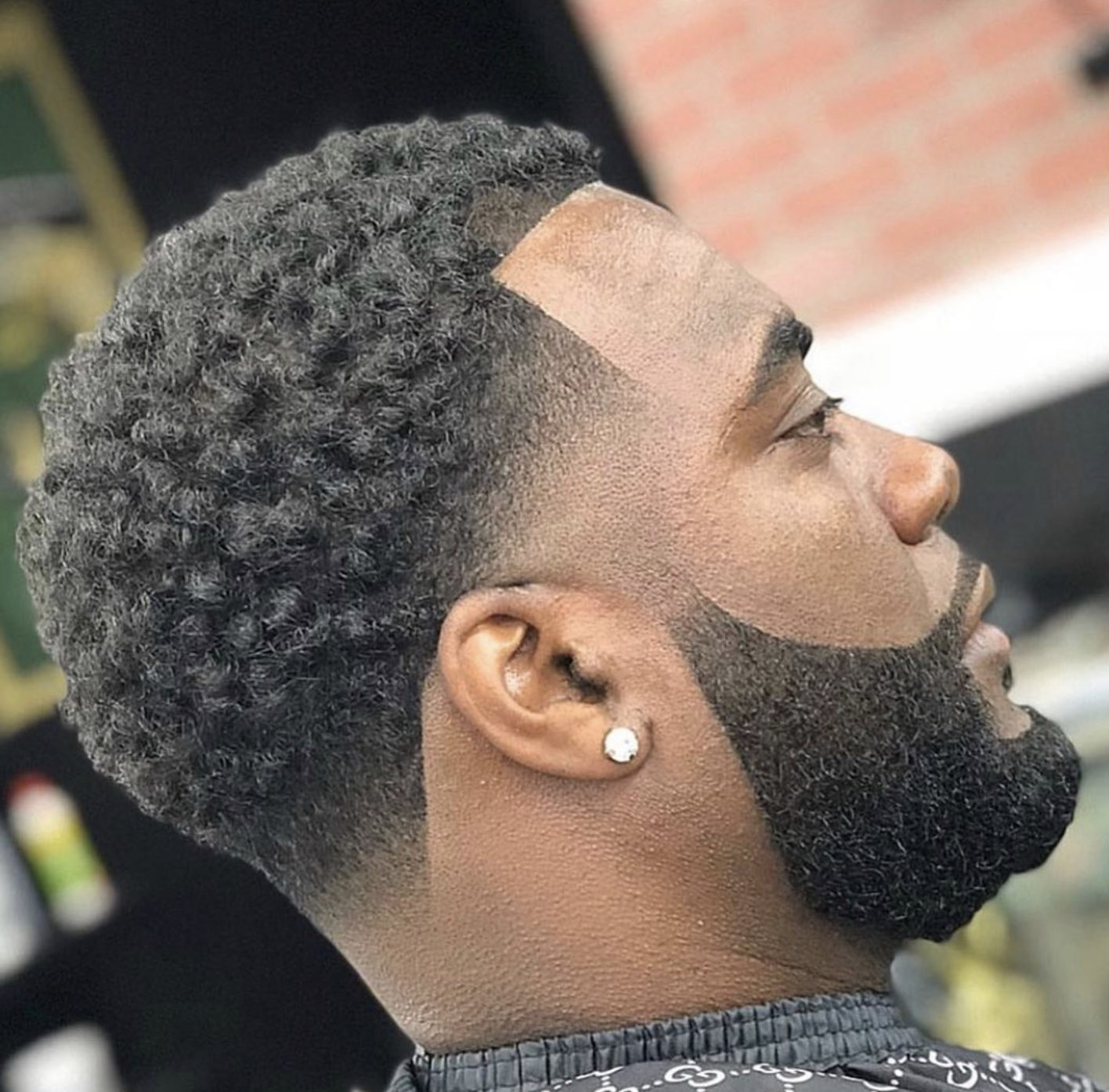 Side profile of a man with a sharp, lined short boxed beard seamlessly faded into a crisp mid-skin fade haircut with textured curls, showcasing precise grooming.