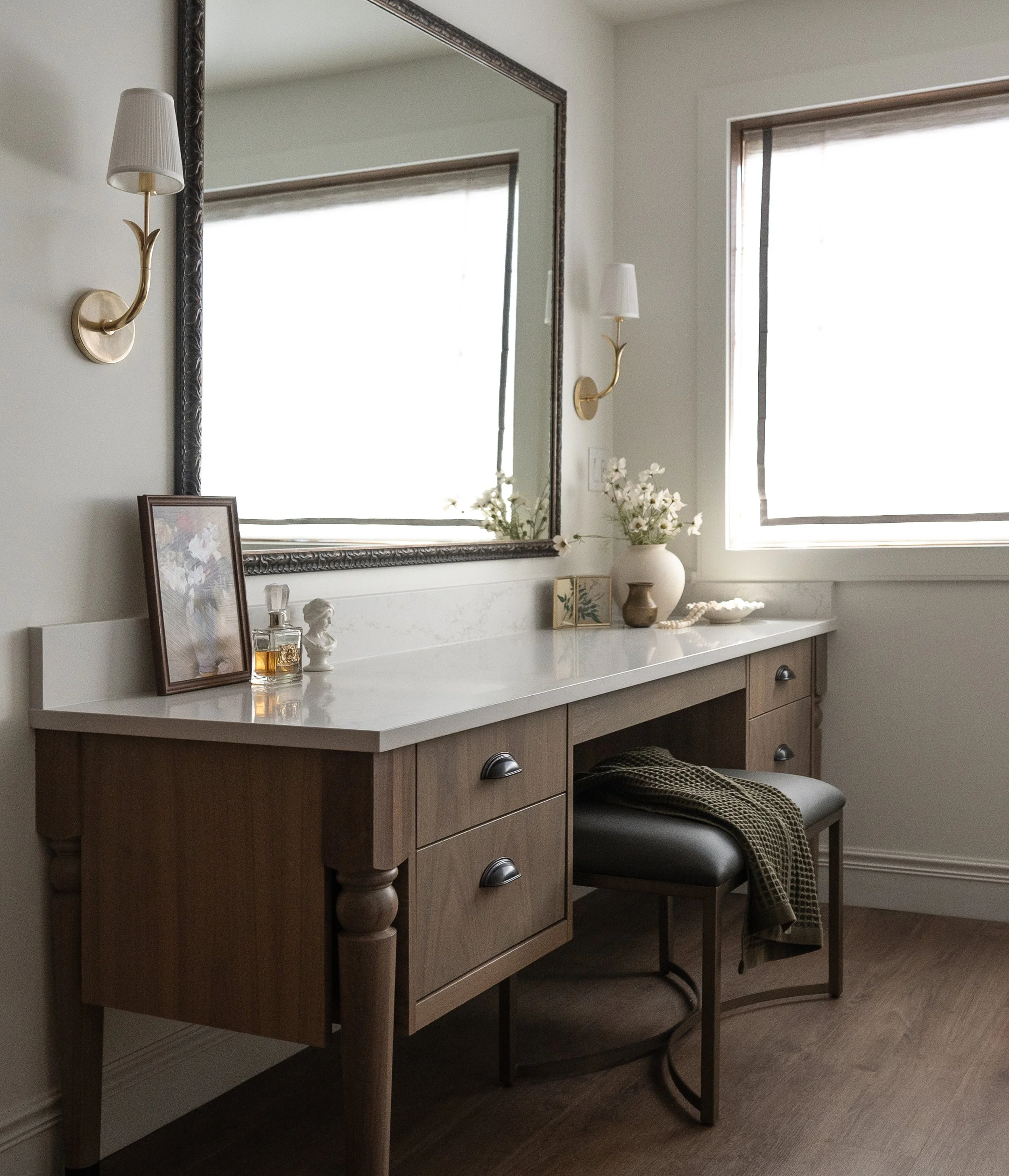 A bathroom vanity with a marble countertop, decorative items, and a large mirror with wall sconces on either side. A stool with a blanket is in front of the vanity, next to a window with blinds.