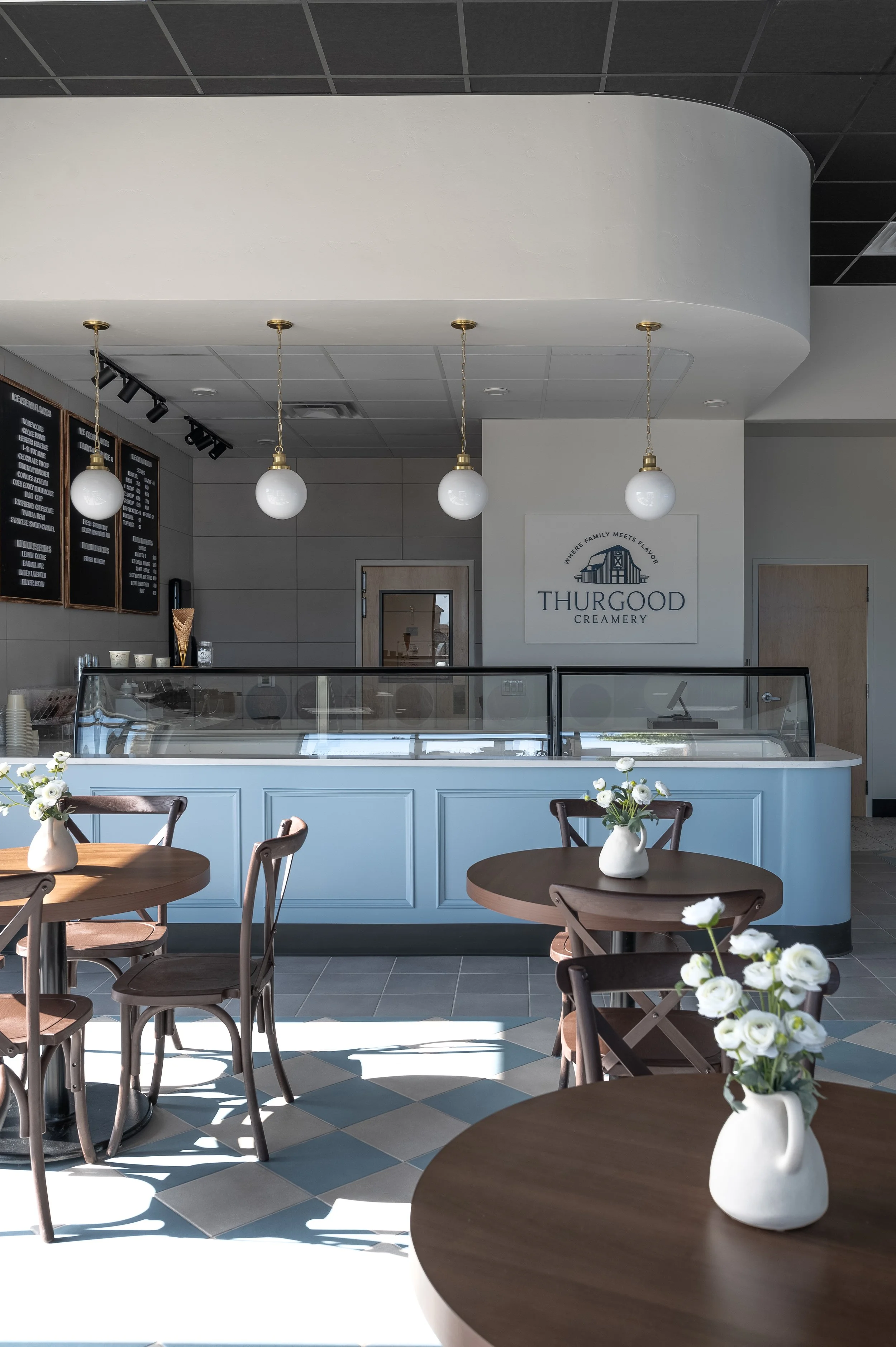 Interior of an ice cream shop with tables and chairs decorated with white flower vases, counter with ice cream display, and a wall sign reading 'Thurgood Creamery'.