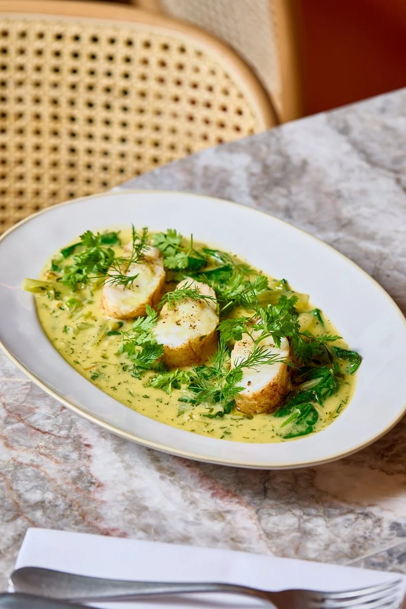 Plated dish with sliced fish and green herbs in a creamy yellow sauce.