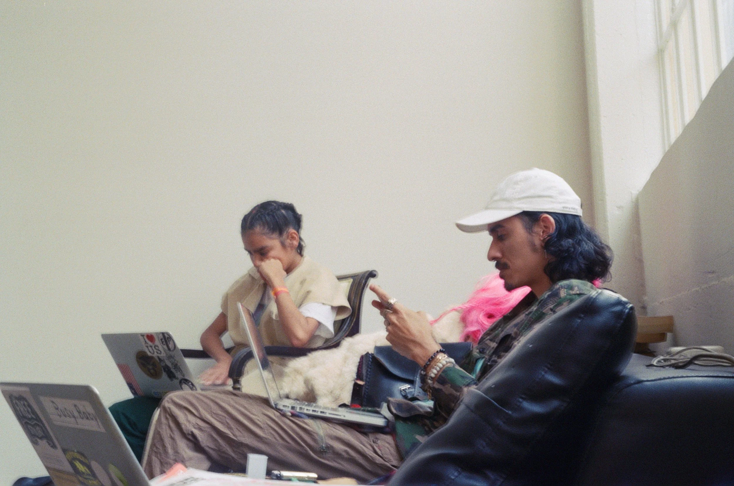 Two young people sitting on chairs, using laptops and a phone in a waiting area with a plain wall and window behind them.