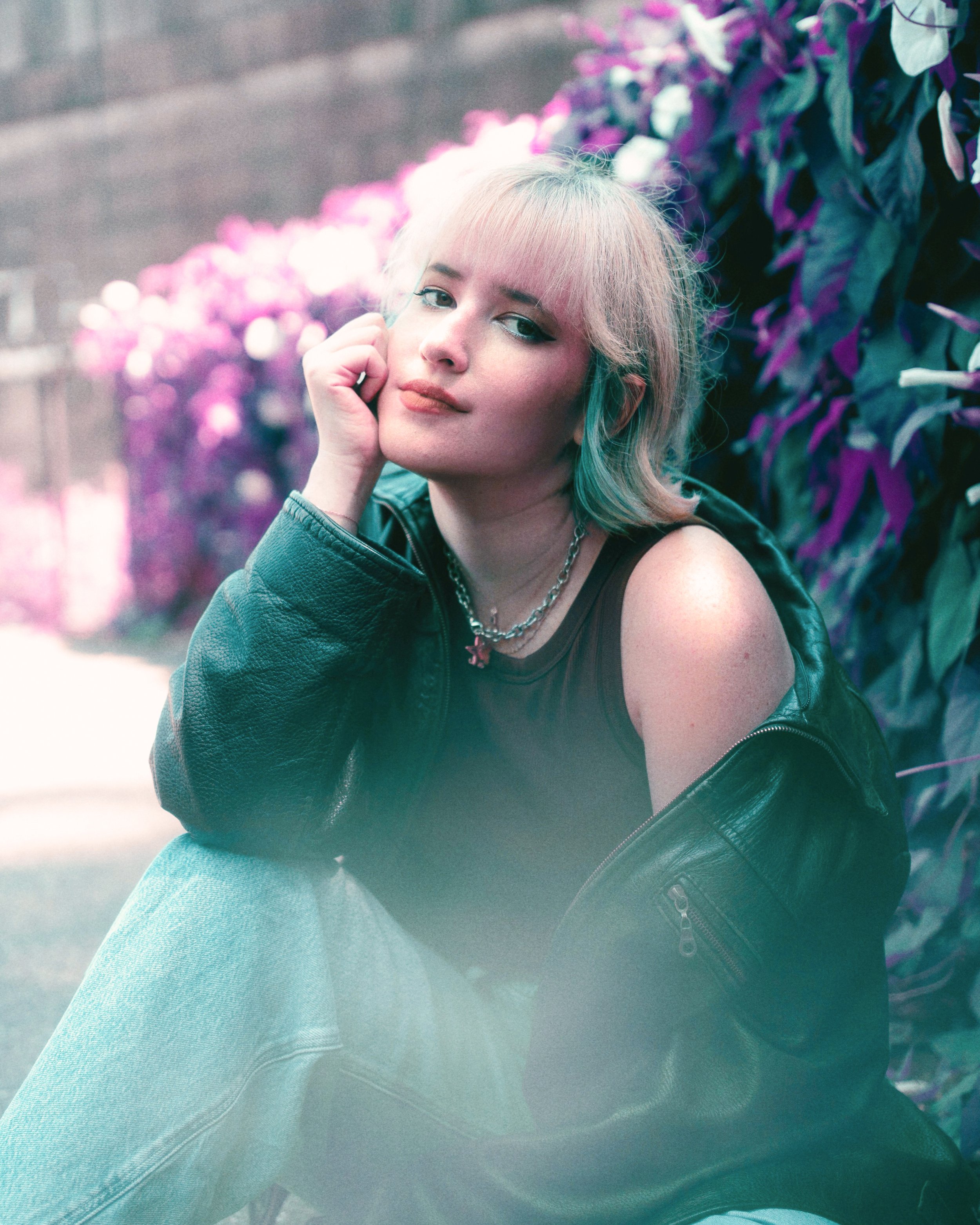 A young woman with blonde hair and blue eyes, dressed in a black tank top and a black leather jacket, sitting outdoors near purple and green plants, resting her face on her hand and looking at the camera with a slight smile.