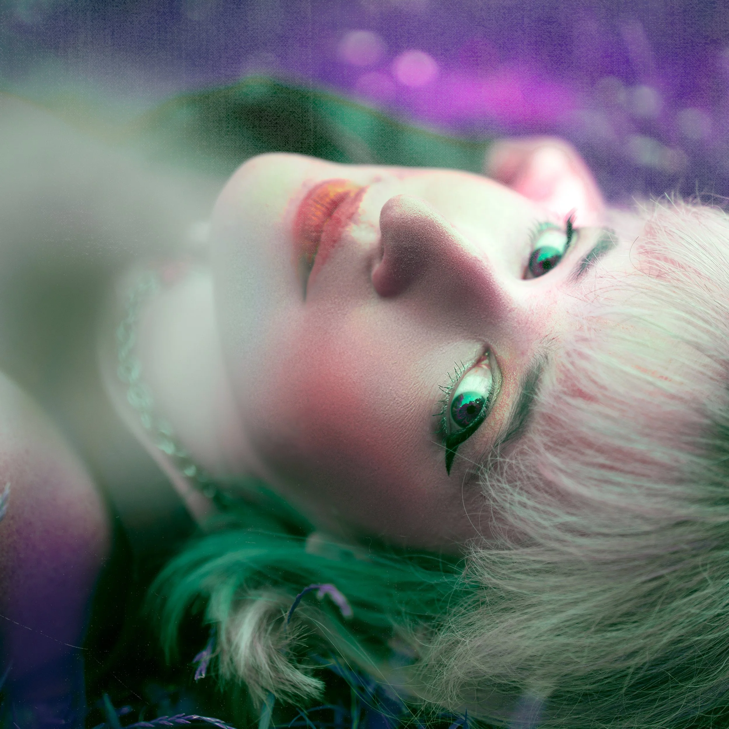Close-up of a person lying down staring sheepishly at a camera, surrounded by colorful, soft-focused lights and grass textures.