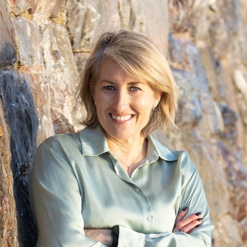 A smiling woman with short blonde hair wearing a silky, light-colored button-up shirt, standing against a textured stone wall with arms crossed.