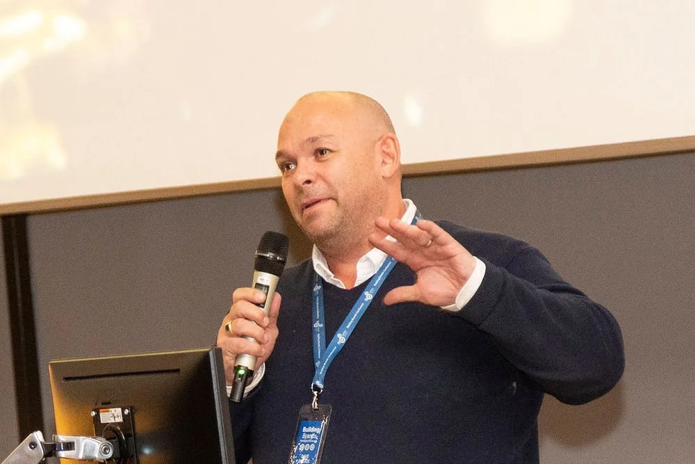 Man speaking into a microphone during a presentation, gesturing with his right hand, with a badge around his neck.