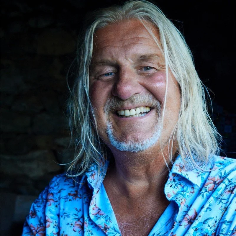 A smiling older man with long gray hair and a beard, wearing a blue floral shirt.