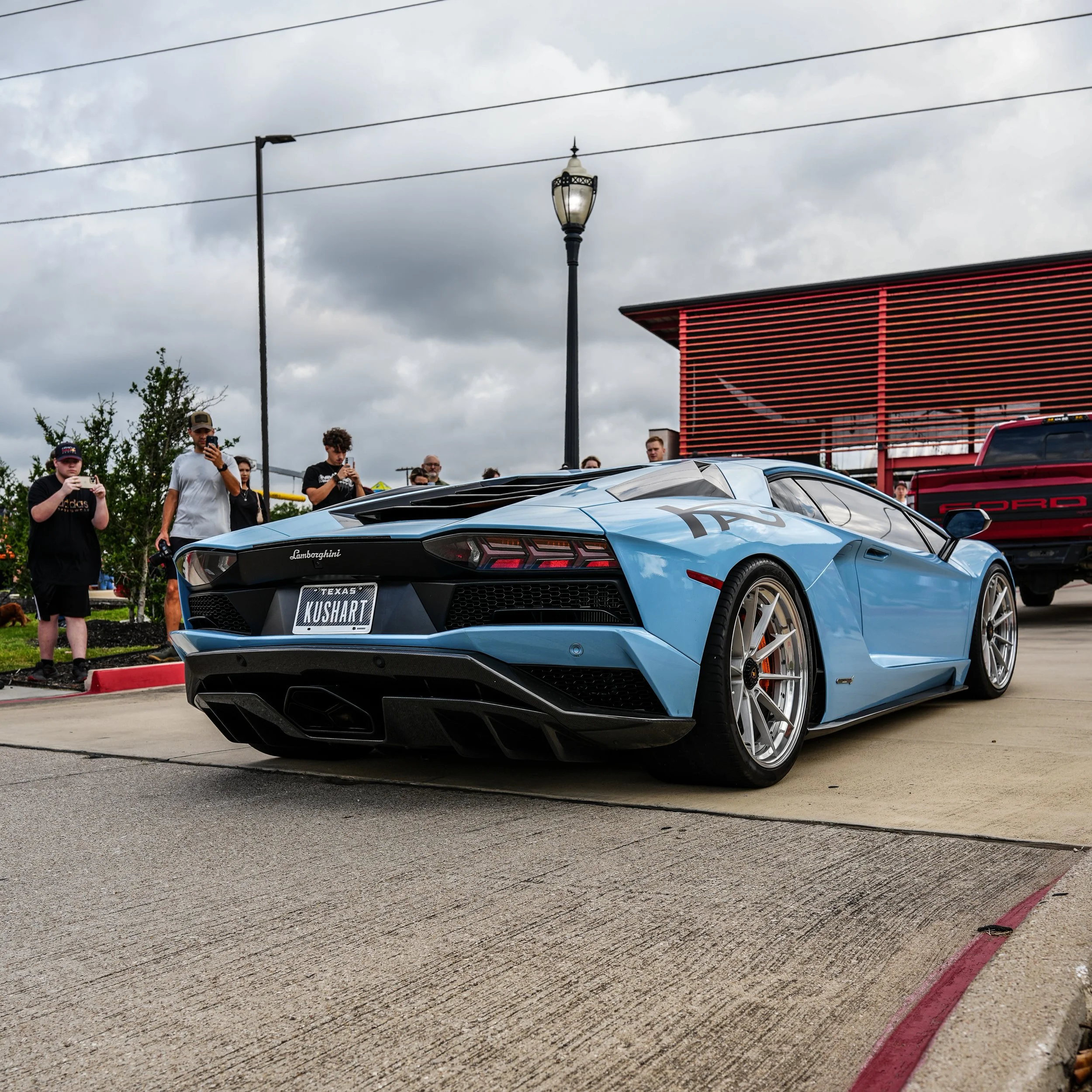 A blue Lamborghini Huracan with a Texas license plate reading 'KUSHART' parked at an outdoor car event, with onlookers taking photos in the background.