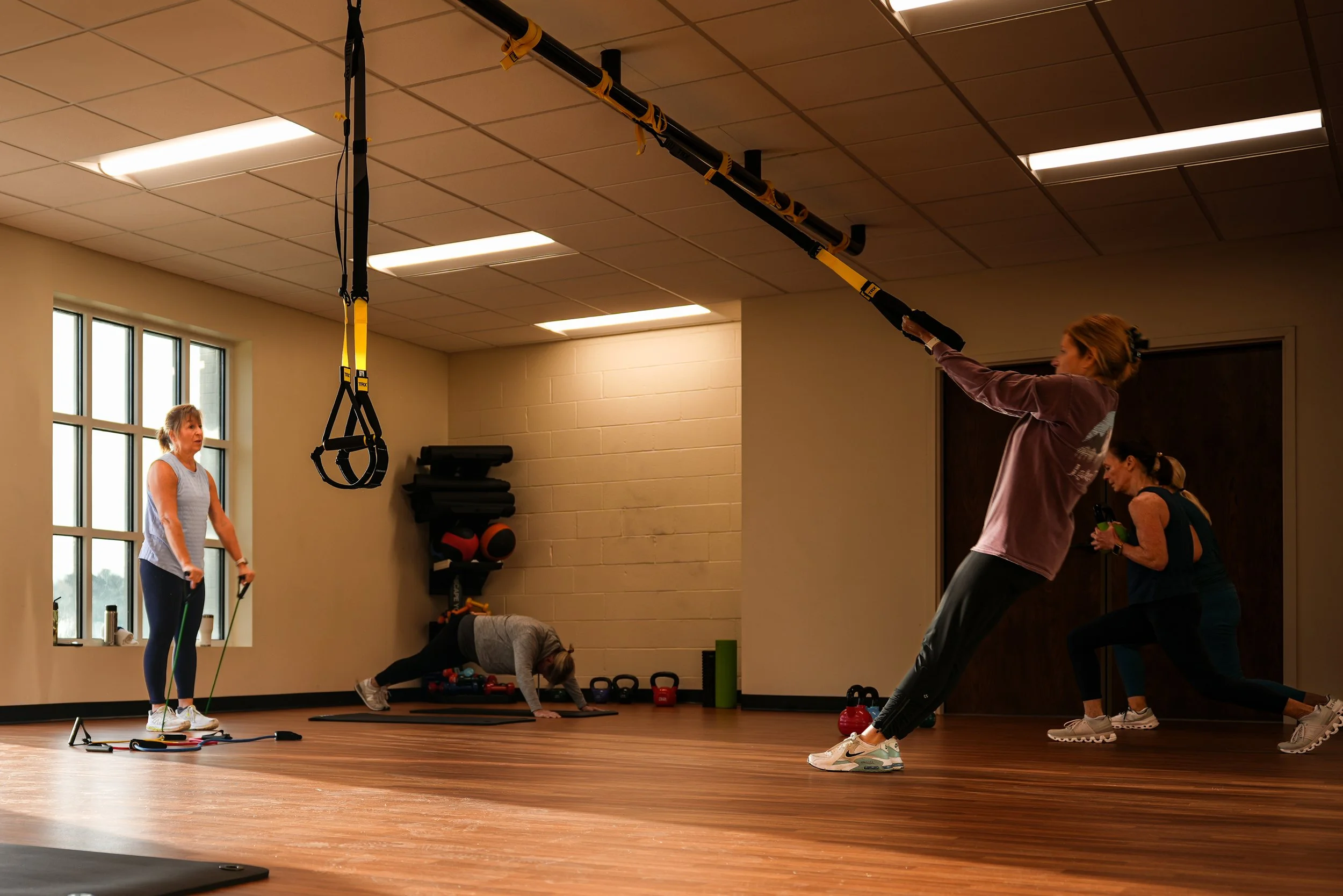 Four women exercise in a gym, using resistance bands, TRX suspension straps, and performing yoga or stretching exercises.