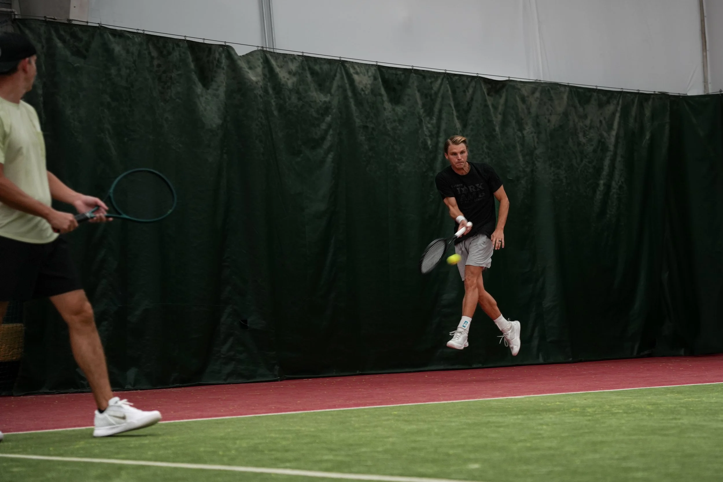 A man in gray shorts and a black shirt is hitting a tennis ball against a green curtain in an indoor tennis court with red and green flooring, while another man in a yellow shirt and black shorts holding a tennis racket watches.