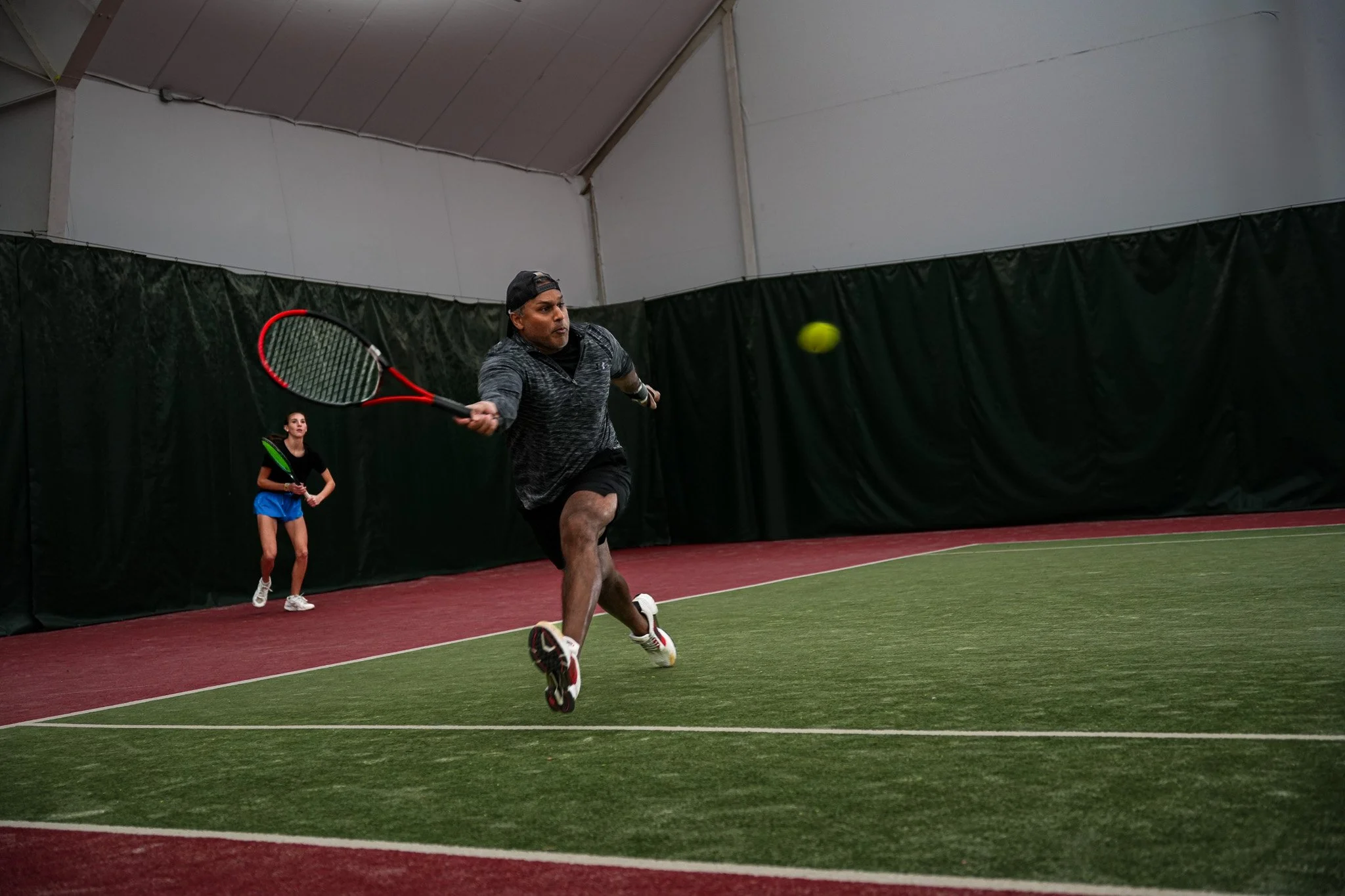 Two people playing indoor tennis, with one man reaching to hit a tennis ball with a racquet, in an indoor tennis court with artificial turf.