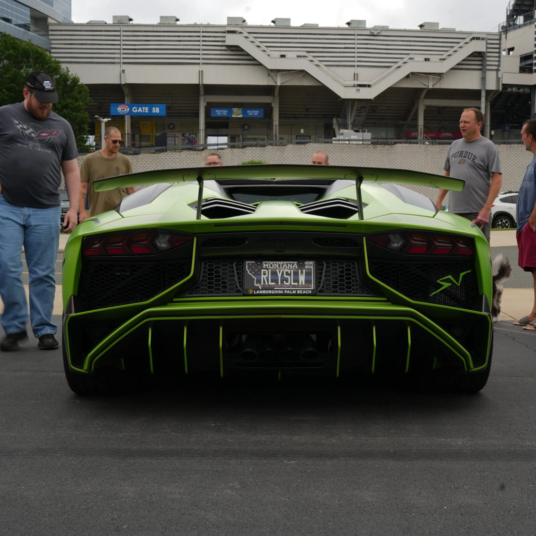 Rear view of a bright green Lamborghini sports car with a spoiler, parked with people standing around it at an outdoor event. The license plate reads 'RLSWL' and mentions Montana and Lamborghini Palm Beach.