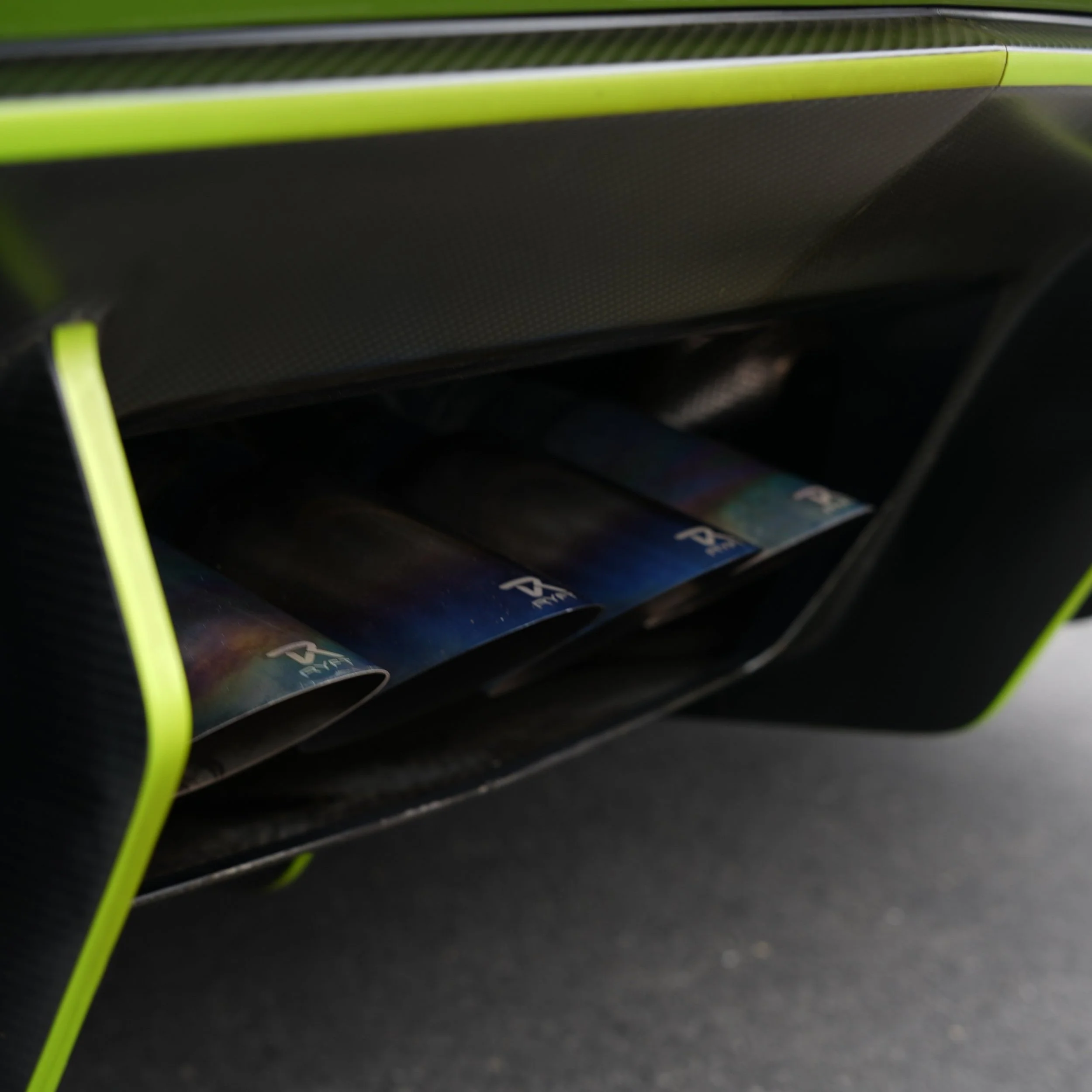 Close-up of a car's exhaust pipe with three chrome tips, part of a black and green vehicle.